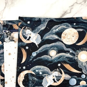 Peut inclure: Une pochette en tissu avec un fond bleu marine foncé et un imprimé céleste composé de lunes, soleils, étoiles et personnages assis sur des croissants de lune. La pochette possède une fermeture éclair bleu foncé avec une breloque feuille en métal argenté.