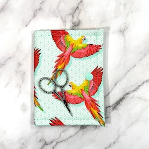 May include: A fabric scissor case featuring a parrot design in red, yellow, and green. The case is light blue with a geometric pattern and a mint green button closure. Silver scissors are visible inside.