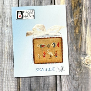 May include: A cross-stitch pattern titled "Seaside Frill" depicting a seaside scene with birds and figures. The design is on a light blue card with the Heart in Hand Needleart logo, tied with a white ribbon.