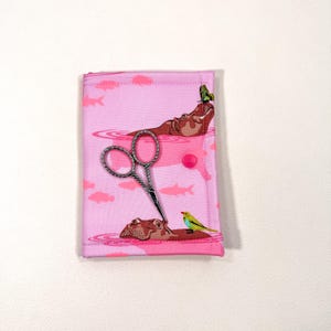 May include: A pink fabric scissor case with a whimsical hippo and fish pattern. The case features a silver pair of scissors and a green butterfly and yellow bird design. The case is closed with a pink button.