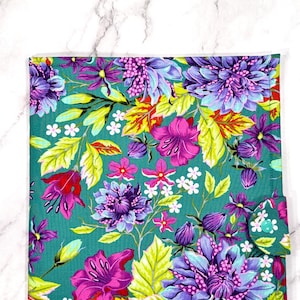 May include: A teal fabric with a vibrant floral pattern in purple, pink, yellow, and white. The design showcases large flowers and leaves. A small flap with a button closure is on the right side.