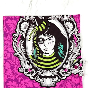 May include: A pink fabric item with a floral pattern. It features a framed illustration of a person with an eye patch, a pirate hat, and green and black striped clothing. The frame is black and white.