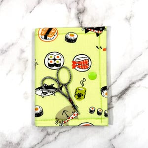 Peut inclure: Un étui de couture en tissu vert clair avec un motif de sushis, représentant divers rouleaux de sushis et illustrations de poissons. Une paire de ciseaux argentés repose sur l'étui, fermé par un bouton-pression vert.