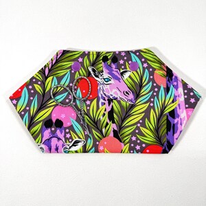 May include: A hexagonal fabric pincushion with a giraffe print. The design features a purple giraffe, green leaves, and pink and red circles on a grey background. A pair of silver scissors rests on the pincushion.