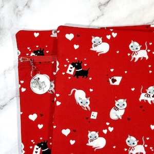 May include: A red fabric pouch featuring a repeating pattern of white cats, black dogs, and white hearts. The pouch includes a red zip closure with a circular metal charm attached, which displays an engraved paw print and leaf design.
