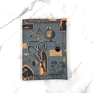 May include: A gray fabric scissor case with a black button closure, featuring a vintage-style print of globes, moths, and architectural elements. Silver scissors rest on the case. The case is on a white marble surface.