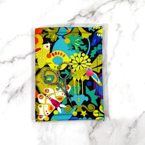 Peut inclure: Ciseaux de broderie argentés posés sur un étui en tissu coloré. L'étui présente un motif vibrant de papillons, de fleurs et de formes abstraites en jaune, bleu et rouge. Une épingle rose ferme l'étui.
