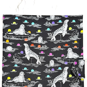 May include: Black fabric featuring a repeating pattern of white seals with black spots, on ice floes and in water. The design includes colourful circles and wave details. A button closure is visible on the right side.