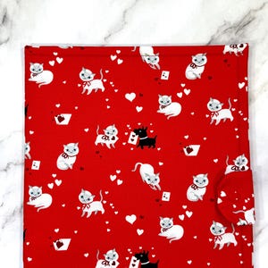 May include: A rectangular red fabric pouch featuring a repeating pattern of white cats with red bows, small black dogs, white hearts, and playing cards. The pouch includes a rounded flap closure on the right side secured with a snap button.