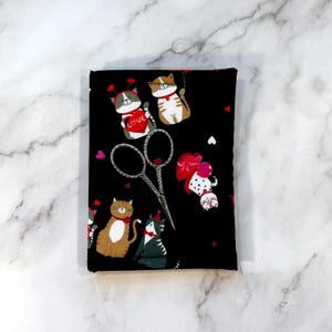 May include: A black fabric featuring a repeating pattern of cartoon cats in various poses, some holding red hearts with the word LOVE printed on them, alongside small red and white heart shapes. A pair of silver metal embroidery scissors is placed on top.