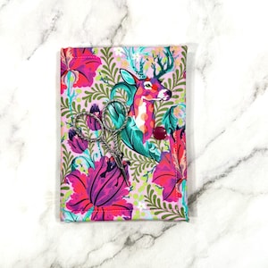 May include: A floral fabric scissor case featuring a deer illustration in pink, teal, and magenta. The case is open, revealing a pair of silver scissors. A red button secures the case. The background is a light pink.