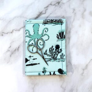 May include: A light blue fabric case featuring a black octopus and sea life print, secured with a single black button. A pair of silver-toned metal embroidery scissors with an intricate handle design rests on top of the fabric case.