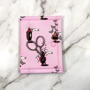 May include: A pink fabric scissor case featuring a pattern of butterflies, lamps, and banners. Silver scissors are placed on the case, which fastens with a pink button. The case has a metal loop for hanging.