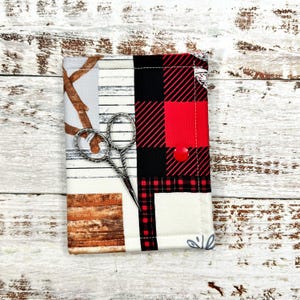 Peut inclure: Une petite pochette en tissu avec un motif à carreaux rouge et noir, un motif en bois et un fond blanc. La pochette a une fermeture à bouton et une paire de ciseaux argentés à l'intérieur.