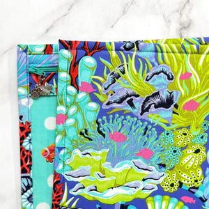 May include: A fabric pouch featuring a vibrant underwater coral reef print with shades of blue, lime green, and red. The design includes stylised sea anemones, coral branches, and small fish. A teal zip with a silver paw print charm is visible.