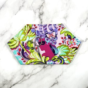 May include: A hexagonal needlework case with a floral and silhouette design. The case features a pink background with a woman's silhouette and a colorful floral pattern. A pair of small scissors is inside the case, secured by two purple snaps.