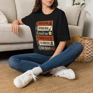 Puede incluir: Una camiseta negra de manga corta con un estampado gráfico de una pila de seis cintas VHS. Las cintas están etiquetadas como Pumpkin Patch Panic, Witchwood Manor, The Midnight Hour, Haunted Hollow, Full Moon Fever y Bone Orchard con iconos.