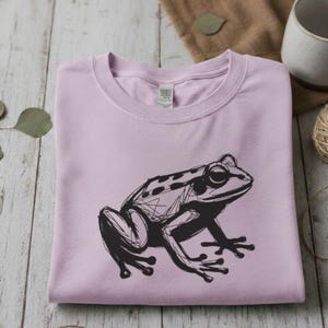May include: A light purple sweatshirt featuring a black sketch of a frog. The frog is drawn in a detailed, artistic style, with visible lines and shading. The sweatshirt is folded neatly on a wooden surface.