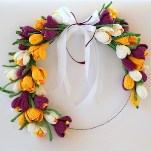May include: A handmade crochet wreath featuring colorful tulips in shades of yellow, white, and purple. The tulips are arranged on a wire frame and tied with a white ribbon. The wreath is a decorative item.