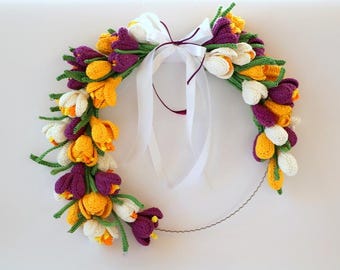 May include: A circular wreath featuring crocheted flowers in shades of purple, yellow, and white with green stems and leaves. A white satin ribbon is tied in a bow at the top center, and a thin, wavy metal wire forms the base of the structure.