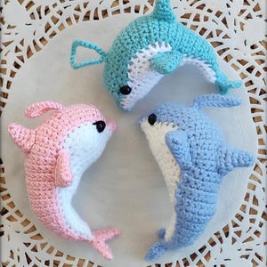 Puede incluir: Tres delfines de ganchillo hechos a mano en tonos azules y rosas, con vientres blancos y ojos negros. Los delfines están dispuestos sobre una superficie blanca y calada. Estos juguetes blandos son perfectos para una guardería o como regalo único.
