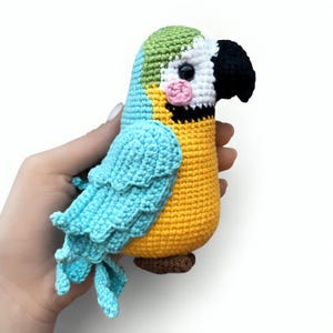 May include: A handmade crochet parrot in bright colors. The bird features a blue body and wings, a yellow chest, and a green head. It has a black beak, white face, and pink cheek. The parrot is a whimsical, handcrafted item.