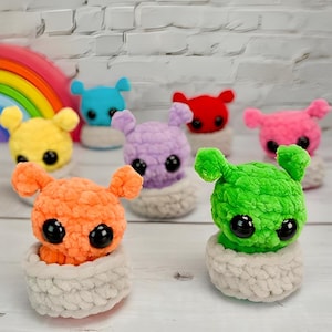 May include: A collection of colorful, handmade crochet creatures. Each creature has large, black eyes and small ears. The creatures are in shades of orange, green, purple, yellow, blue, red, and pink. They sit in small, off-white crochet baskets.
