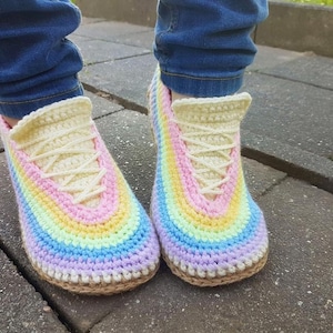 Puede incluir: Un par de zapatillas de ganchillo hechas a mano con un esquema de colores arcoíris. Las zapatillas presentan un diseño similar a una zapatilla deportiva con una lengüeta y cordones de color crema y una suela marrón. Las rayas coloridas incluyen rosa, amarillo, azul y morado.