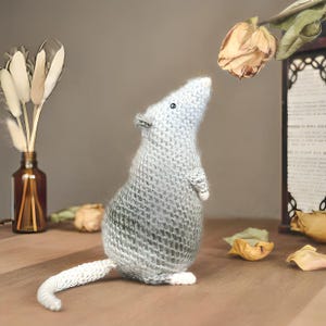 Puede incluir: Un juguete de rata tejido a mano, de color gris claro, con una larga cola blanca. La rata está mirando una rosa seca. El juguete tiene pequeños ojos negros y pequeñas patas blancas. El fondo incluye una botella de vidrio marrón con flores secas.