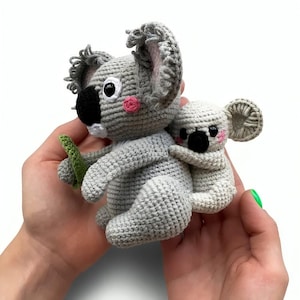 May include: Two handmade crochet koala bears. The larger koala is gray with a black nose, pink cheeks, and a green leaf. A smaller koala is hugging the larger one. The koalas are held in a person's hands.