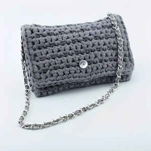 Könnte beinhalten: Eine graue gehäkelte Clutch mit einem silbernen Kettenriemen. Die Tasche hat ein strukturiertes Stichmuster und einen klaren, funkelnden Edelsteinverschluss. Der Kettenriemen ist eine Kombination aus silbernem Metall und grauem Stoff.