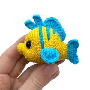 May include: A crocheted yellow and blue fish with black eyes. The fish has blue fins and stripes, and is held in a hand against a white background. The fish is a handmade item.