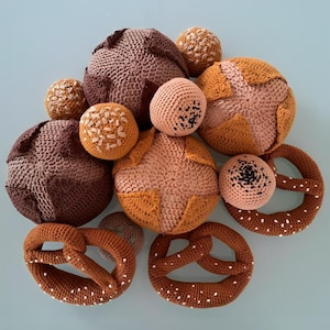 Op de afbeelding: Een verzameling gehaakte voedselartikelen, waaronder broden, broodjes en pretzels. De items zijn in tinten bruin, bruin en beige, met witte accenten op de pretzels. De opstelling staat op een lichtblauwe ondergrond.