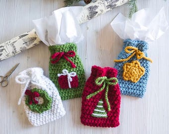 Könnte beinhalten: Vier handgefertigte gehäkelte Geschenktüten in Weiß, Grün, Rot und Blau. Jede Tasche hat ein einzigartiges Feiertagsdesign, darunter ein Kranz, ein Geschenk, ein Weihnachtsbaum und ein Stern. Die Taschen sind mit weißem Seidenpapier gefüllt und mit passendem Garn gebunden.