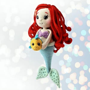 Pode incluir: Boneca de sereia de crochê com cabelo ruivo, top roxo e cauda azul. A boneca segura um peixe amarelo. A cauda da sereia é azul claro com detalhes verdes. Feito à mão.