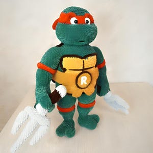 May include: A green plush toy of a Teenage Mutant Ninja Turtle, featuring a red mask and orange accents. The turtle has a yellow chest with a white "R" and holds a white trident and a brown and white weapon. The toy is designed to resemble a cartoon character.