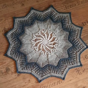 Puede incluir: Un tapete de ganchillo hecho a mano en forma de estrella, en tonos grises y azules. El intrincado diseño presenta un patrón de espiral central y un borde festoneado. El tapete se muestra sobre una superficie de madera.