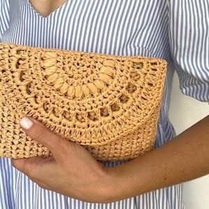 May include: A light tan woven straw clutch bag with a flap closure. The front of the bag has a decorative, semi-circular pattern. The bag is held against a blue and white striped shirt.