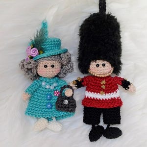 May include: Two crocheted dolls: one in a teal dress and hat with a pearl necklace and a black purse, and the other in a red jacket and black pants, resembling a British Royal Guard. Both dolls have black eyes and are on a white surface.