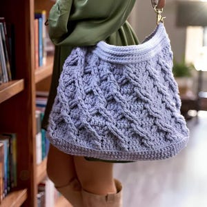 May include: A light purple, handmade crochet bag with a textured cable knit design. The bag has a brown leather strap and a gold-colored clasp. The bag is held by a person wearing a green dress and tan boots. The bag is approximately 14 inches wide.