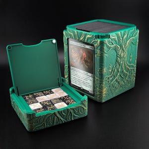 May include: A green deck box with a textured tree relief and gold accents, featuring an open compartment containing black and white dice with numerical markings and a display slot holding an Ashaya, Soul of the Wild trading card.