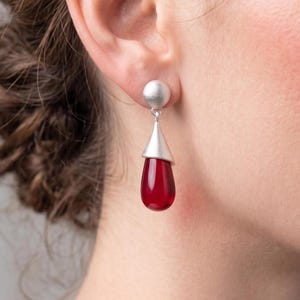 Peut inclure: Boucles d'oreilles argentées avec un clou rond et un pendentif en forme de goutte d'eau en pierre précieuse rouge foncé. Les boucles d'oreilles ont un design moderne et minimaliste, contrastant le métal poli et la pierre rouge vibrante.