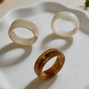 Può includere: Tre anelli in resina in varie tonalità di marrone e crema sono su un vassoio bianco con bordi ondulati. Gli anelli hanno un design grosso e arrotondato, con un effetto marmorizzato. Gli anelli sono un accessorio moda.