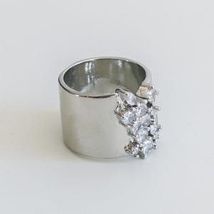 Puede incluir: Un anillo ancho plateado con un grupo de piedras claras y brillantes. El anillo tiene un acabado liso y pulido y un diseño moderno. Las piedras están dispuestas en un patrón floral o de estrella. El ancho del anillo es de aproximadamente 1,5 cm.