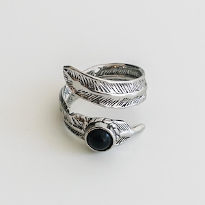 Puede incluir: Un anillo plateado con diseño de pluma, que se enrolla alrededor del dedo. El anillo presenta una pequeña piedra negra redonda. El anillo está sobre un fondo blanco liso.