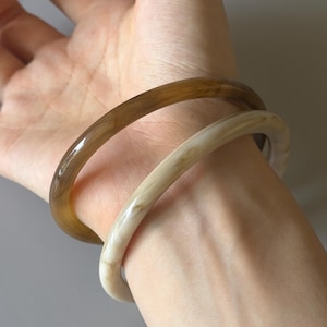 Op de afbeelding: Twee doorschijnende armbanden, één lichtbruin en de andere gebroken wit, worden om een pols gedragen. De armbanden zijn glad en afgerond, met een glanzende afwerking. De lichtbruine armband ligt bovenop de gebroken witte armband.