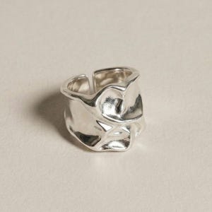May include: A silver ring with a distinctive, textured design. The ring has an irregular, organic shape, with a slightly open band. The surface reflects light, creating highlights and shadows. The ring appears to be made of metal.