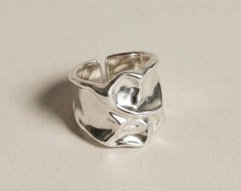 May include: A silver-colored ring with a unique, textured design. The ring has an irregular, organic shape, with a slightly open band. The surface of the ring reflects light, creating highlights and shadows. The ring appears to be made of metal.