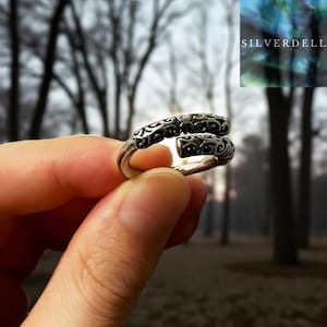 Peut inclure: Bague en argent avec un design ouvert et des motifs de volutes complexes. La bague est présentée sur un fond flou d'arbres. Le mot "SILVERDELL" est visible dans le coin supérieur droit.