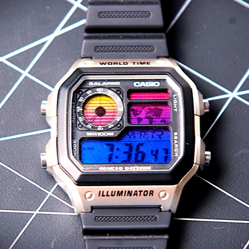May include: A digital Casio watch with a black resin strap and silver-tone case. The watch face displays the time in blue, with world time and alarm features. The face has a colorful sunset-like design.